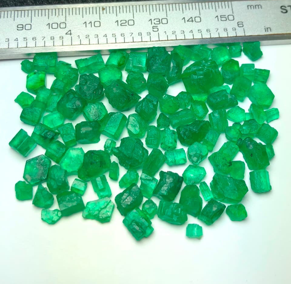 A pile of small, rough-cut green emerald-cut gemstones displayed on a white surface with a metal ruler at the top for scale.