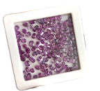 A hand holding a small square white tray filled with many faceted purple gemstones, photographed close up against a softly blurred background.